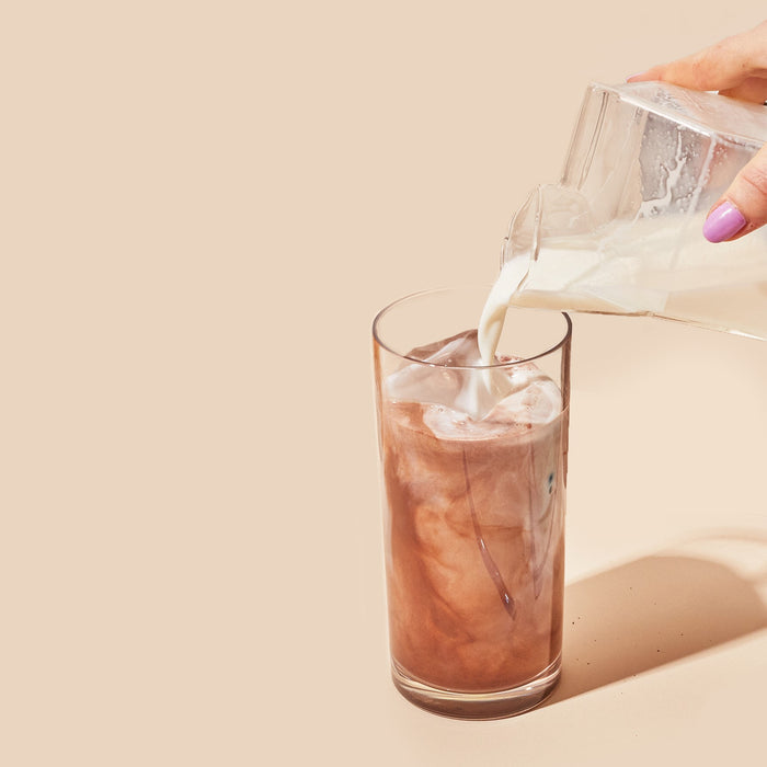 A hand with purple nail polish pours milk from a glass pitcher into a tall glass of iced coffee mixed with blume Organic Reishi Hot Cacao Latte Powder, creating swirling patterns against a solid light beige background.