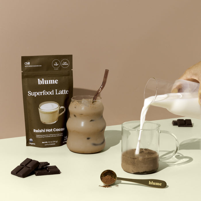 A hand pours milk into a glass with blume Organic Reishi Hot Cacao Latte Powder. Nearby are an iced hot chocolate, a blume latte packet, superfood powder, chocolate pieces, and a spoon of powder on a beige surface.
