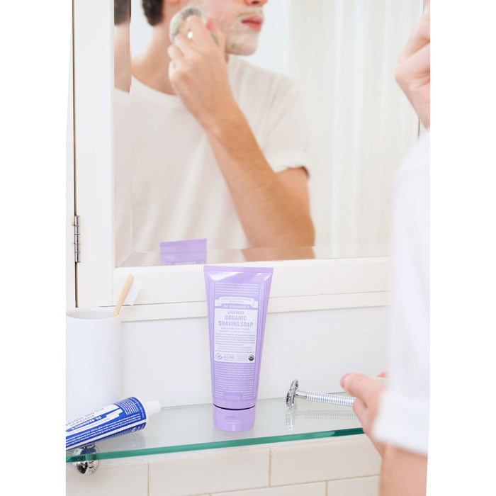 A person shaves in front of the bathroom mirror. On a glass shelf sits Dr. Bronners Organic Shaving Soap in a lavender tube, alongside a razor and a blue cream, creating a bright, clean scene.