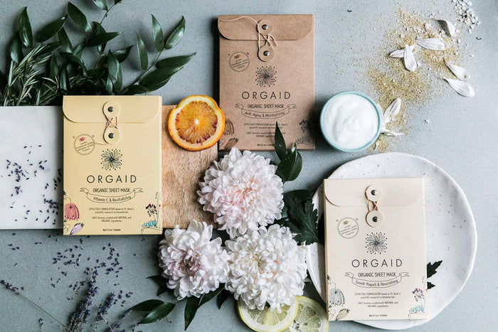An ORGAID Organic Sheet Mask Multi-Pack is showcased in a flat lay arrangement with Ecoderma fabric packaging, surrounded by flowers, lavender, citrus slices, cream, and petals, beautifully highlighting a natural skincare theme.