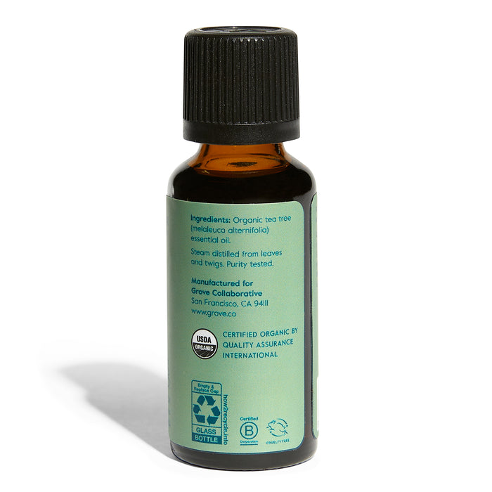 A small brown glass bottle with a green label displays text and logos, listing organic tea tree essential oil from Grove Co. for purifying aromatherapy. It includes USDA Organic certification and a recycling symbol.