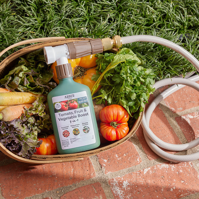 An Organic Tomato, Fruit & Vegetables 3-in-1 Boost RTS spray bottle by Arber sits in a vegetable-filled basket with tomatoes and leafy greens, promising a harvest increase. A hose is ready for use, while the basket rests on bricks with lush grass behind.