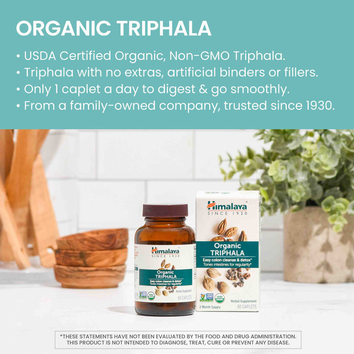 A bottle and box of Himalaya Organic Triphala, sitting on a kitchen counter beside a cutting board with vegetables in the background. The product is USDA Certified Organic and Non-GMO, promoting easy digestion with no additives. Text highlights product details.