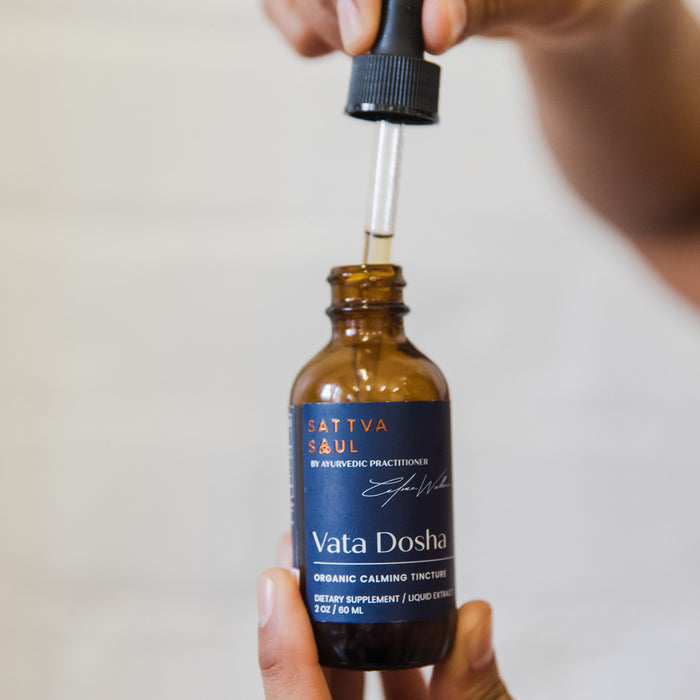A hand holds a dropper above an amber bottle labeled Sattva Soul. The 2 oz Organic Vata Tincture - Calming, infused with Ayurvedic herbs, caters to the Vata Dosha and rests against a softly blurred background.