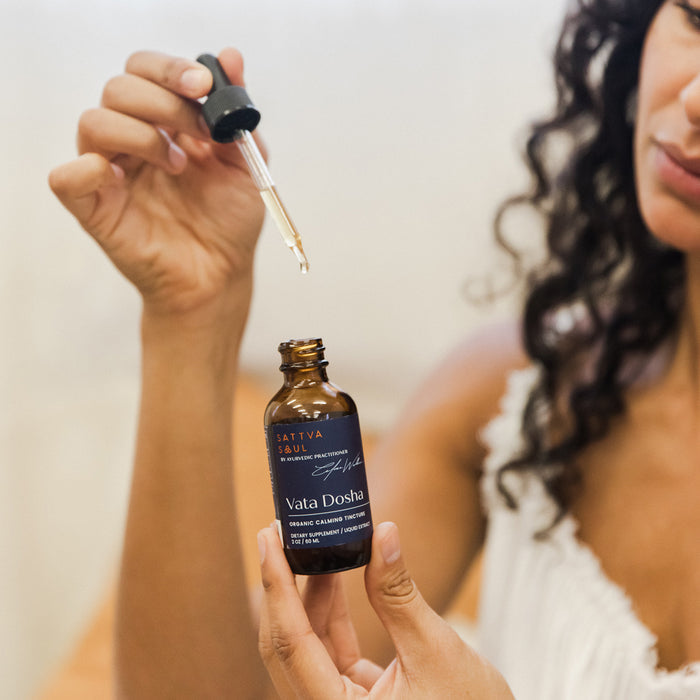 A person wearing a white top holds a dropper above a brown bottle of Organic Vata Tincture - Calming by Sattva Soul. The tincture, enriched with Ayurvedic herbs, sits on a wooden surface, with the persons curly hair partially visible.