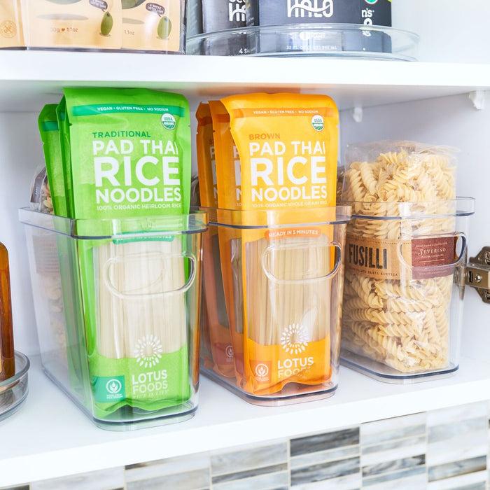 A pantry shelf features an iDesign Organizer Bin with Handles made from recycled plastic, holding two clear containers of Pad Thai rice noodles—one green and one orange—and a third with Fusilli pasta. Jars and boxes complement the neat food display in the background.
