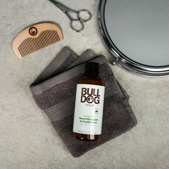 A bottle of Bulldogs Original Beard Shampoo/Conditioner rests on a folded gray towel. Scissors, a wooden comb, and a round mirror are nearby on a light countertop—ideal for those dedicated to organic beard care with vegan formulations.