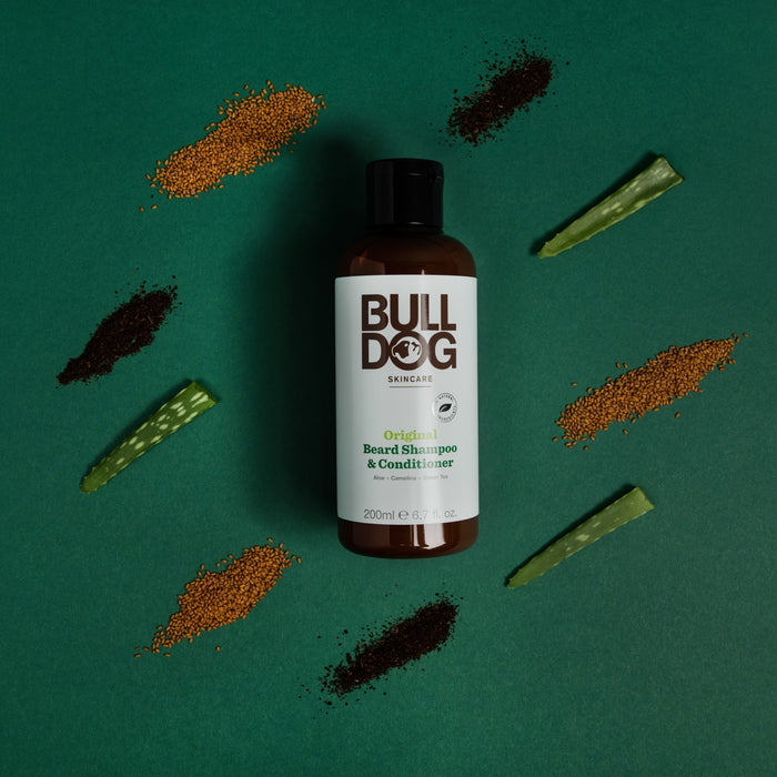 A Bulldog Original Beard Shampoo/Conditioner bottle, an organic beard care essential, is centered on a green backdrop with symmetrical heaps of seeds, coffee grounds, and aloe leaf segments around it.