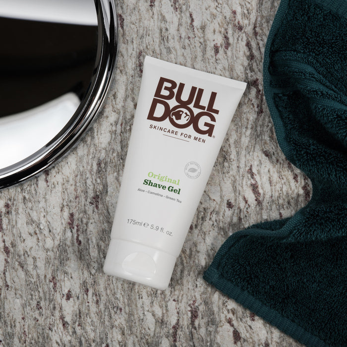 The Bulldog Original Shave Gel, renowned for its natural skincare benefits, sits on a textured surface beside a green towel. The white tube with brown and green text is near a partially visible round mirror in the corner.