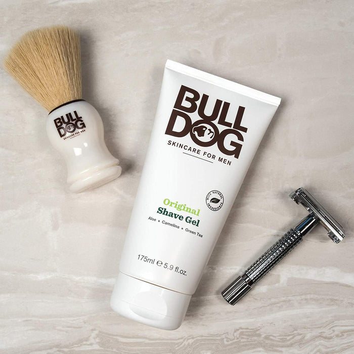A Bulldog Original Shave Gel, celebrated for natural skincare, lies on marble beside a BULL DOG brush and metal razor. The gel features vegan ingredients like aloe, camelina, and green tea.