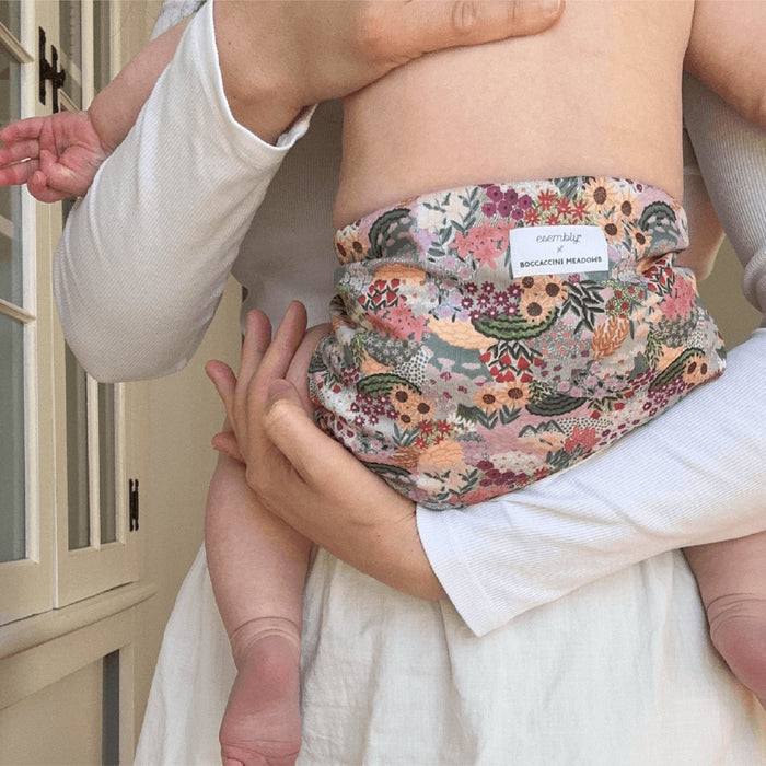 A person cradles a baby in an Esembly Baby Outer Diaper Cover + Swim Diaper with floral patterns, made from 100% OEKO-TEX certified recycled fabric. A white cabinet with glass panels in the background is bathed in soft light, creating a warm and gentle atmosphere.
