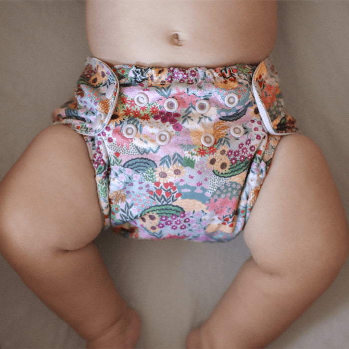 A baby lies on a soft surface wearing an Esembly Baby Outer Diaper Cover + Swim Diaper, Size 2, made from 100% OEKO-TEX certified recycled fabric with a vibrant floral and animal print. The reusable diapers multiple snaps offer adjustable sizing while the babys legs are visible against a blurred background.