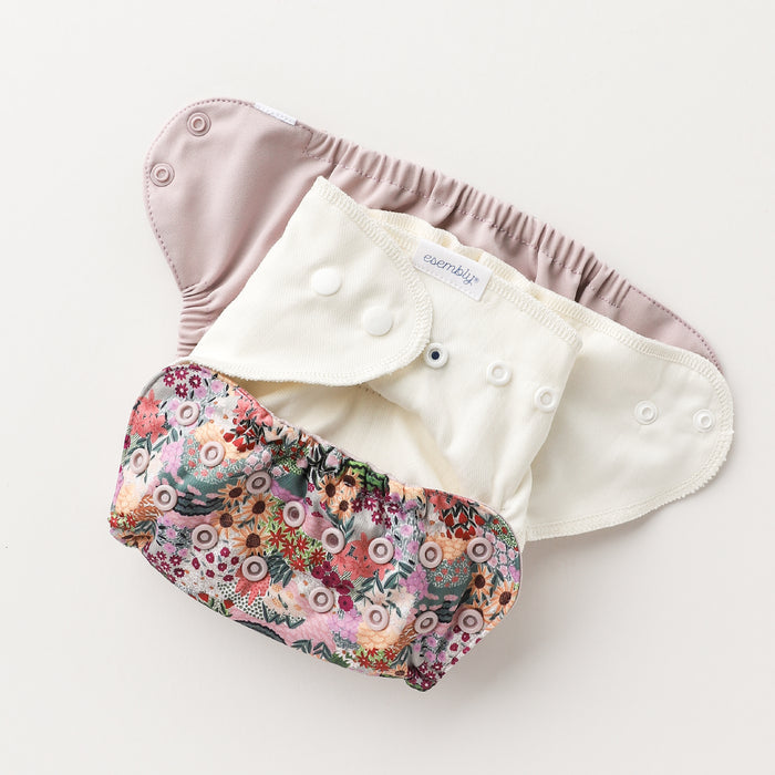 Three Esembly Baby Outer Diaper Cover + Swim Diapers, made from 100% OEKO-TEX certified recycled fabric, are arranged on a simple backdrop: the top in light pink, the center in pristine white, and the bottom with a vibrant floral pattern—all featuring white snaps for easy fastening.