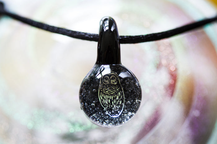 Round borosilicate glass pendant with a holographic foil owl inside and cremation ash, hung on an adjustable cord.