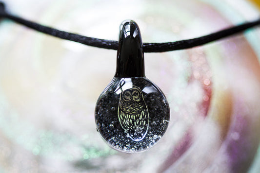 Round borosilicate glass pendant with a holographic foil owl inside and cremation ash, hung on an adjustable cord.