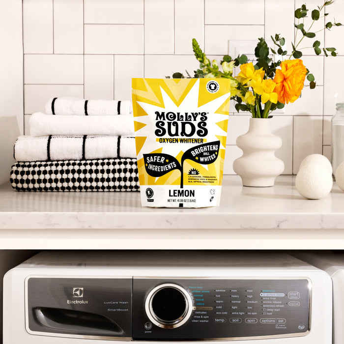 A package of Molly's Suds Oxygen Whitener sits on a white shelf above a washing machine, with folded towels and a vase of yellow and white flowers in the background.