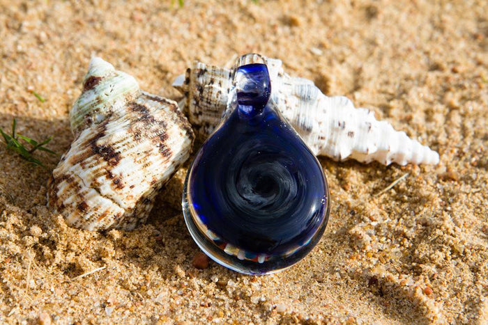 Blue glass reef pendant with swirling pattern sits on sand beside seashells infused with cremains