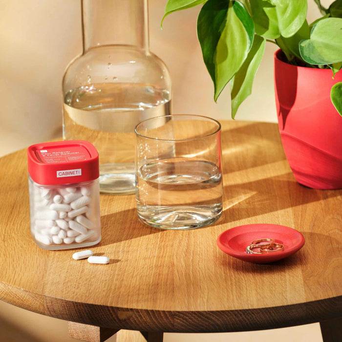 A wooden table holds a bottle and glass of water, a container labeled Pain Reliever & Fever Reducer - Acetaminophen Tablets USP, 500 mg by Cabinet Health, a dish with jewelry, and a plant in a red pot. The warm light casts an inviting atmosphere.