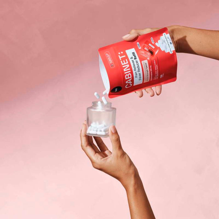 Two hands are seen pouring Pain Reliever & Fever Reducer - Acetaminophen 500 mg tablets from a red refill pouch labeled Cabinet Health into a clear jar, set against a pink background, showcasing its role as a sustainable refill solution.