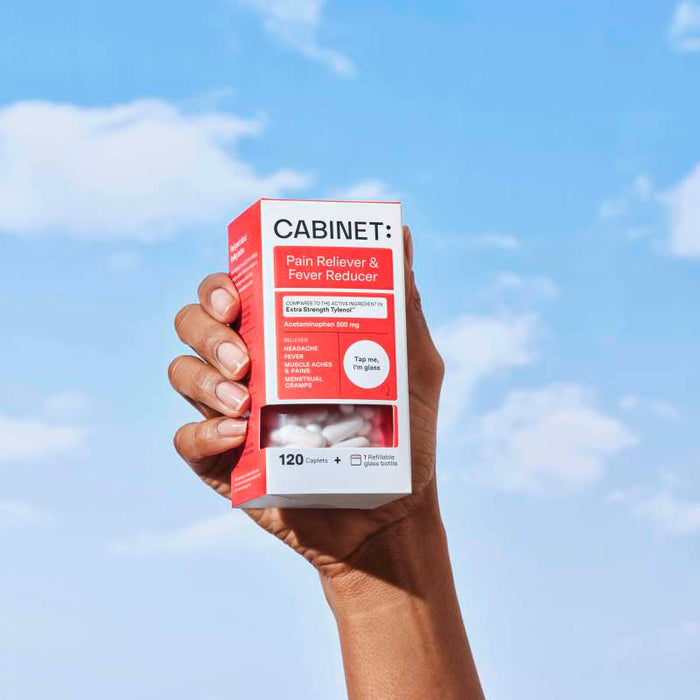 Against a blue sky with clouds, a hand holds a red and white box labeled Cabinet Health: Pain Reliever & Fever Reducer. It contains 120 caplets of sustainable acetaminophen tablets, each 500 mg.