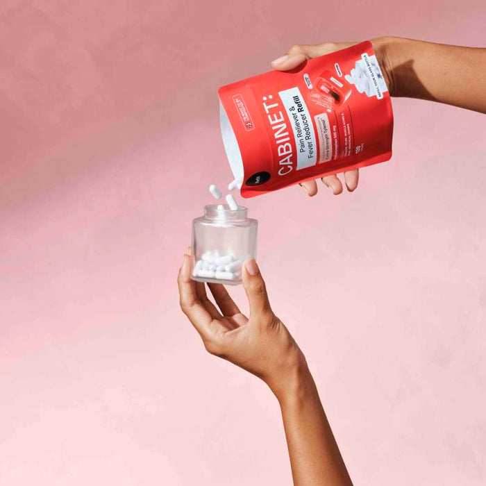 Two hands are shown: one holds a red Cabinet Health pouch, the other a clear jar. White acetaminophen tablets, known for reducing fever, spill from the Pain Reliever & Fever Reducer Refill - Acetaminophen Tablets USP, 500 mg pouch into the jar against a pink background.