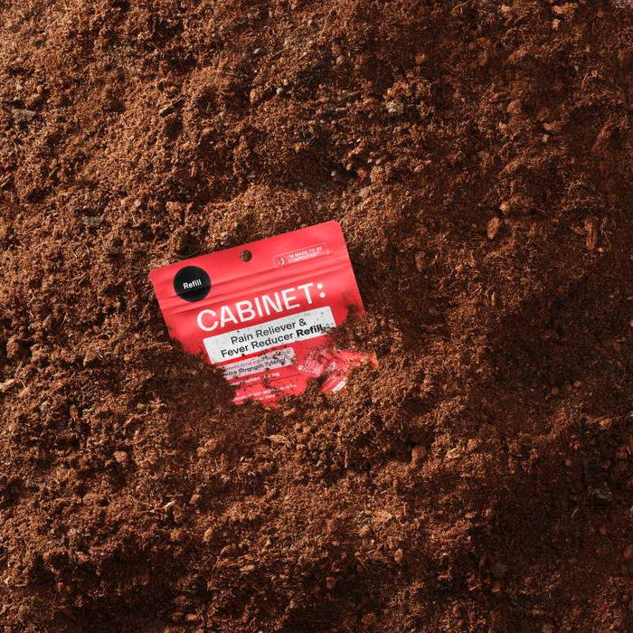 A red packet labeled Cabinet Health: Pain Reliever & Fever Reducer Refill - Acetaminophen Tablets USP, 500 mg sits in lush soil with its top peeking out. This plastic-free packaging blends seamlessly with the earth around it.