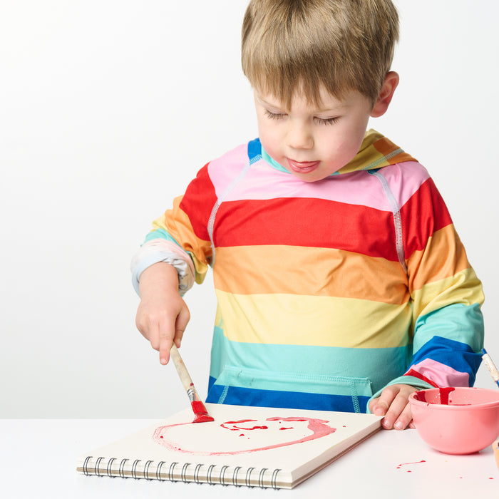 A young child in a colorful striped hoodie uses the Elseware Unplug Paintbrush Set with water-based paints to create a red face in a sketchbook, focusing intently beside a pink bowl of paint on the white table.