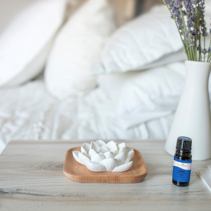 The Passive Lotus Diffuser by Plant Therapy, shaped like a white lotus, sits on a beech wood tray on a light wood table with essential oils nearby. A bed with white pillows and lavender in a vase completes the peaceful scene.