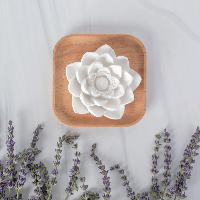 The Passive Lotus Diffuser by Plant Therapy, a white flower-shaped piece infused with essential oils, sits on a natural beech wood square dish. Lavender sprigs lie below on light gray marble, creating a serene ambiance.