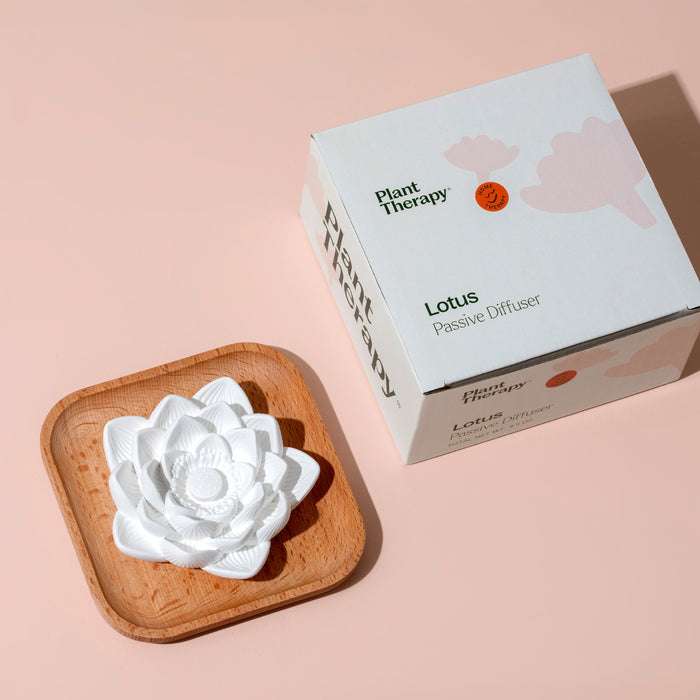 A Passive Lotus Diffuser by Plant Therapy, shaped like a white lotus, sits on a natural beech wood tray next to a box labeled Plant Therapy Lotus Passive Diffuser on a light pink surface. The box shadow reveals the lotus design, suggesting an enchanting journey with essential oils.