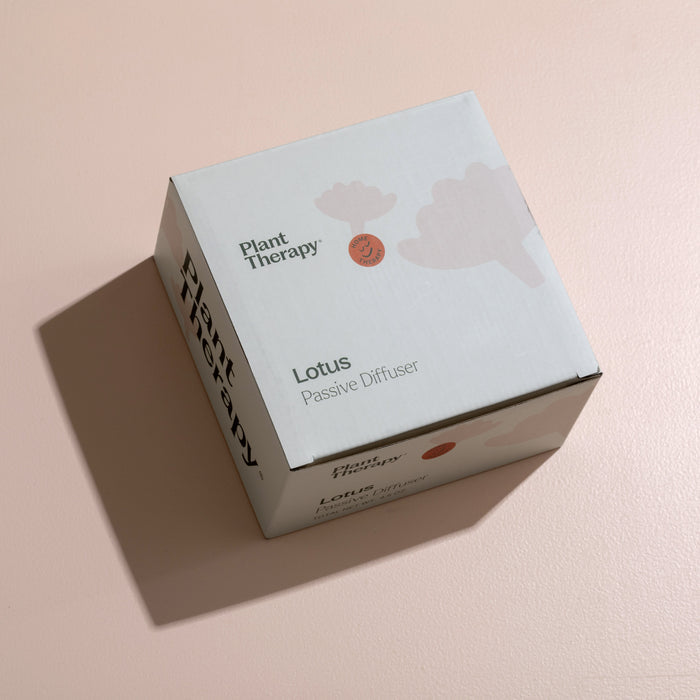 A box labeled Plant Therapy with Passive Lotus Diffuser printed on it rests elegantly on a light pink surface, casting a shadow. The abstract design pairs beautifully with the natural beech wood, enhancing the graceful diffusion of essential oils.