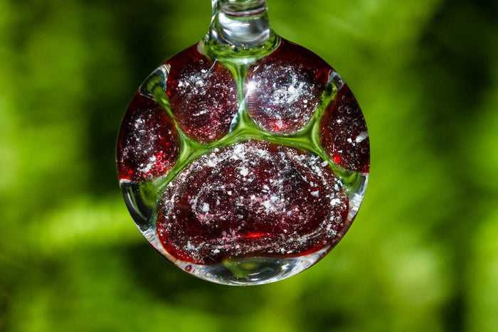 Paw print glass pendant with red hues and white speckles infused with cremains against a green backdrop