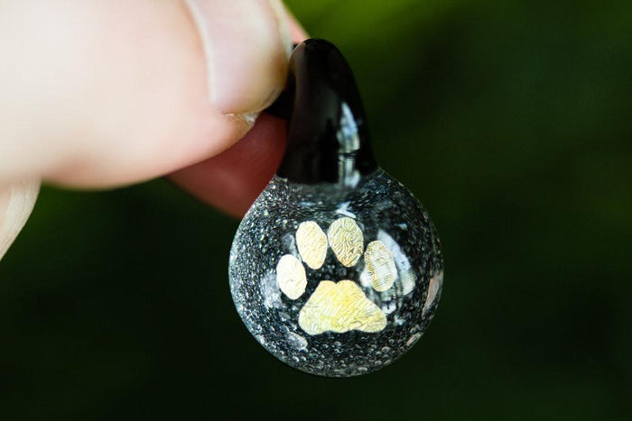 Small glass pendant infused with cremation ashes featuring a gold pawprint inside, hanging from a cord.