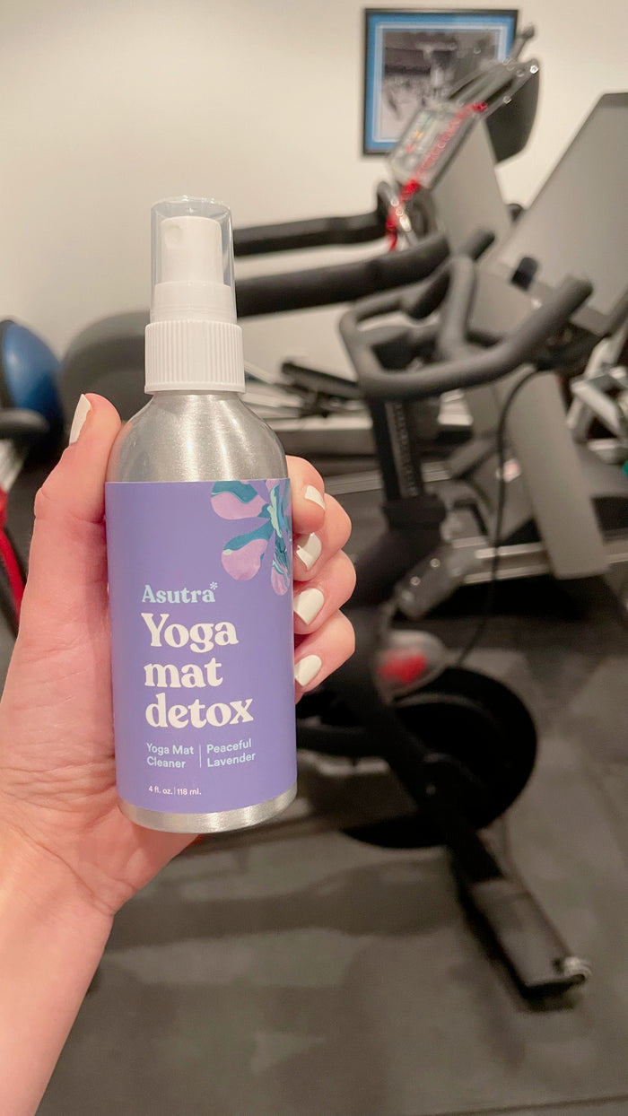 A hand holds a bottle of Asutras Peaceful Lavender Yoga Mat Cleaner, distinguished by its purple label. Infused with lavender essential oil, this cleaner is shown in a gym environment alongside exercise equipment like a stationary bike.