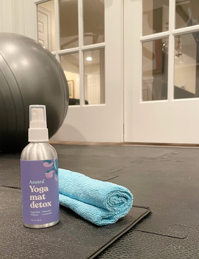 Asutras Peaceful Lavender Yoga Mat Cleaner, accompanied by a blue cloth on a black mat next to an exercise ball, is set against a white-framed glass door backdrop, emphasizing the premium quality of Asutra products.