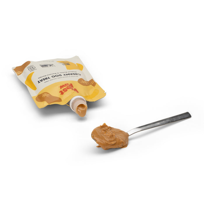 A West Paw squeeze pouch labeled Peanut Butter Banana Creamy Dog Treat lies on a white surface, partially open, spilling some contents onto a stainless steel spoon. The label features peanut illustrations, emphasizing the treats delightful peanut butter flavor.