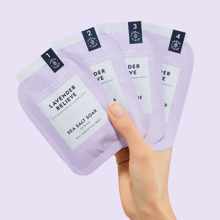 A hand holds four purple packets labeled Pedi Moments, by VOESH New York, reminiscent of a spa experience. Each packet, numbered 1 to 4, is neatly arranged against a pale purple backdrop, evoking the essence of a luxurious pedicure kit.