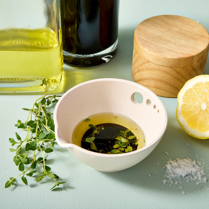 The Peel, Grate & Prep Dish from Modern Sprout beautifully showcases its culinary functions filled with olive oil, balsamic vinegar, and fresh thyme. Nearby are a bottle of olive oil, a wooden container, half-cut lemon peel, and salt flakes on a light green surface.