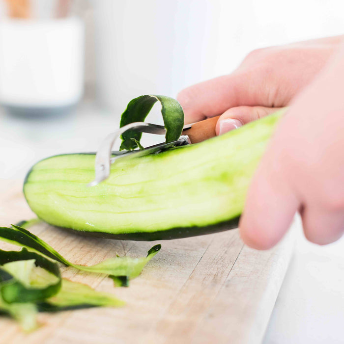 Close-up of a hand using the Full Circle Peeler with a comfortable handle and stainless steel blade to easily peel a cucumber on a wooden cutting board, with peelings gathered neatly beside it.