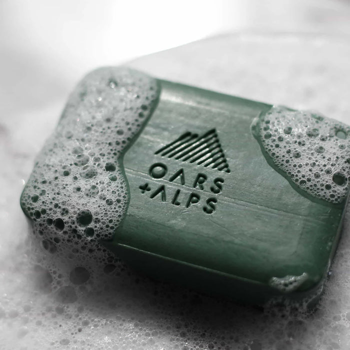 A green Oars + Alps Charcoal Bar Soap with bubbles on its surface rests on a wet, white background. The soap features an embossed mountain logo and the brand name.