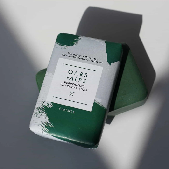 An Oars + Alps Charcoal Bar Soap, featuring exfoliating activated charcoal and partially wrapped in green and white packaging, sits atop an unwrapped green soap bar on a white surface with shadows.