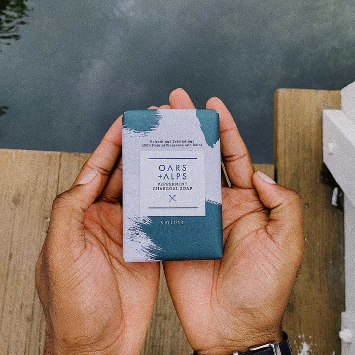 A person holds an Oars + Alps Charcoal Bar Soap, infused with peppermint oil and activated charcoal, over a wooden dock by water. The teal and white packaging is visible.
