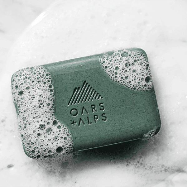 The Oars + Alps Charcoal Bar Soap, a green exfoliating bar with white lather and bubbles, sits on a wet, white surface displaying the brand’s logo and name.