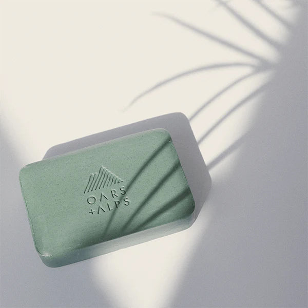 A green rectangular Charcoal Bar Soap by Oars + Alps, embossed with a mountain logo and set on a white surface. A plant’s shadow highlights its refreshing peppermint oil formula.