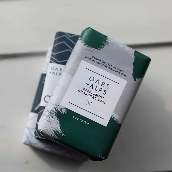 Two Oars + Alps Charcoal Bar Soaps, featuring peppermint oil and activated charcoal in green and white packaging, are stacked on a geometric-patterned bar and set on a white surface.