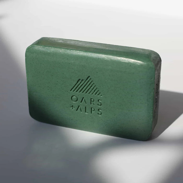 A green rectangular Charcoal Bar Soap by Oars + Alps, embossed with the brand’s logo, sits on a white surface with subtle shadows; it’s infused with Activated Charcoal.