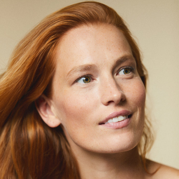 A person with long red hair smiles gently, looking off to the side, their lips subtly accented by the moisturizing touch of Kari Grans Peppermint Lip Whip Treatment Balm. The background is softly blurred, adding a warm tone to the image.