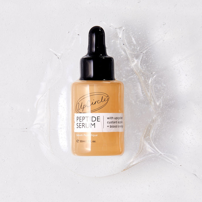 UpCircles Peptide Serum with Custard Apple + Blood Orange, a 30 ml bottle with a black dropper lid, rests on a white surface amid a clear splash. The label showcases upcycled custard apple + blood orange, enhancing collagen effortlessly.