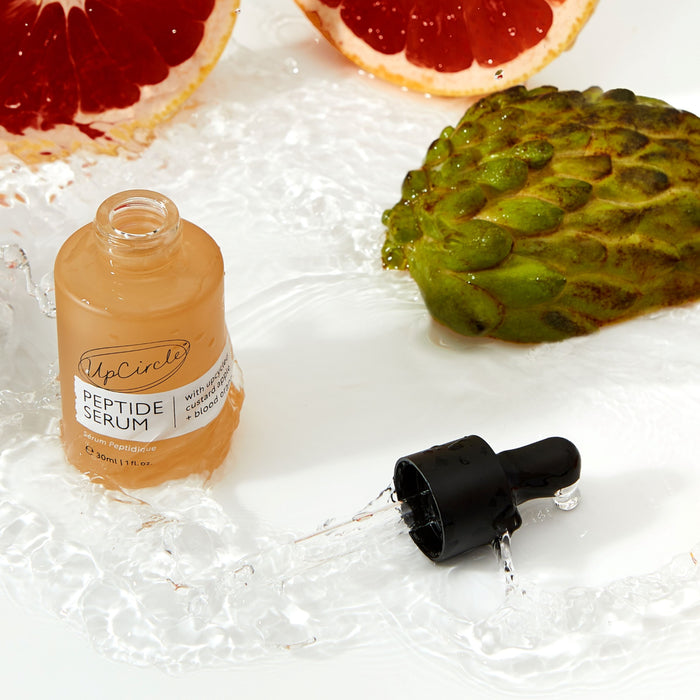 An UpCircle Peptide Serum with Custard Apple + Blood Orange bottle lies on a wet surface alongside half a red grapefruit and custard apple, embodying freshness and natural ingredients. This skincare powerhouse enhances collagen production.