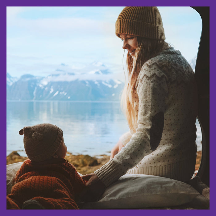 A woman and a child in knitted sweaters and hats relax by a camper van, taking in the calm lake and snow-capped mountains. With New Chapters Perfect Postnatal™ Multivitamin for energy, mood & lactation support, they embrace not only peace but mindful wellness on this tranquil retreat.
